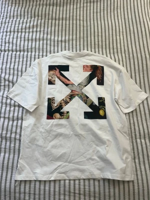 Vit Off-White t-shirt med tryck - Jätte snygg vit t-shirt med svart logga och handmotiv på bröstet. Baksidan har stort grafiskt tryck med konstmotiv i Off-Whites ikoniska pilar. Klassisk passform och rund halsringning. Perfekt för dig som gillar streetwea. Den är stor och passar L-XL