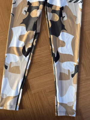Camo träningsleggings från Gavelo - Träningsleggings från Gavelo med camomönster i beige, svart, vitt och brunt. Tillverkade av 73% återvunnen polyester och 27% spandex för maximal stretch och komfort. Tight passform och hög midja, perfekta för gymmet eller löprundan.