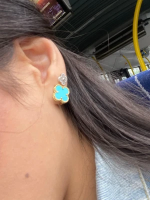 Blå klöverformade örhängen guldpläterad hög kvalitet - Snygga örhängen i klöverform med en stor blå detalj omgiven av guldfärgade pärlor och en mindre silverfärgad blomma ovanför. Perfekta för att ge din look en färgklick och unik touch. Passar dig som gillar trendiga accessoarer med lite extra bling.