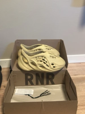 Yeezy Foam Runner sandfärgade tofflor - Säljer ett par Yeezy Foam Runner i sandfärg. Tofflorna har en futuristisk design med många ventilationshål och är tillverkade i ett lätt, skummaterial som ger skön komfort. Perfekta för dig som vill ha något unikt och trendigt på fötterna.