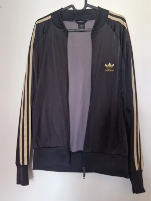 Adidas kofta  - Säljer en svart Adidas kofta med klassiska guldiga ränder längs ärmarna och Adidas logga på bröstet. Dragkedja framtill, ribbade muddar och två fickor. Perfekt för en sportig och retro look. Finns en liten prick fläck framtill som jag inte provat att ta bort.