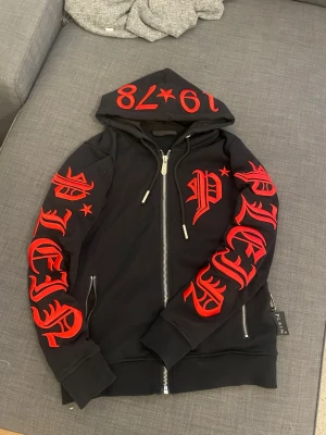  Philipp Plein zip up  - Säljer en svart hoodie från Philipp Plein med dragkedja och stora röda tryck i gotisk stil på ärmar och huva. Huva med snörning och siffrorna '1978' tryckta ovanpå. Två fickor med dragkedja och ribbade muddar. Märkeslapp på sidan. Ganska ovanligt jätte fint skick och tags medföljer. 