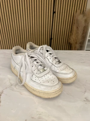 Vita Nike Air Force 1 sneakers - Klassiska vita Nike Air Force 1 sneakers i läder med perforerad tå, vita skosnören och robust sula. Ikonisk design med Nike swoosh på sidan och metallbricka med 'AF1' på snörningen. Perfekta för en streetwear-look.