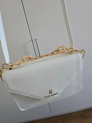 Vit axelväska från Steve Madden - Vit Steve Madden väskan i modell BINDIO. Väskan har magnetstängning och justerbar axelrem. Jag  fick väskan som en gåva för ett år sedan, har dock inte använt den då jag sällan bär vitt. Valde "mycket bra skick" då den fått små defekter av att ha förvarats trångt i garderoben.