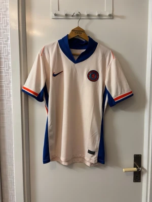 Chelsea FC  matchtröja Nike - Snygg retro Chelsea FC matchtröja från Nike i vitt med blå och röda detaljer. Tröjan har korta ärmar, V-ringad krage och klubbmärke på bröstet. Tillverkad i lätt och ventilerande material, perfekt för fotboll eller streetwear.