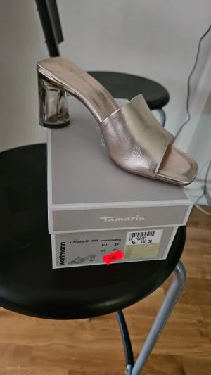 Metallic pumps med blockklack Tamaris - Snygga metallicfärgade pumps från Tamaris med öppen tå och bred rem över foten. Skon har en cool transparent blockklack som ger en modern touch. Perfekta för dig som vill sticka ut med stilrena detaljer och trendig design. Kvitto finns 