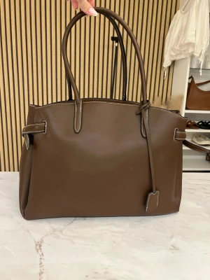 Brun handväska i läderlook - Stilren brun handväska med dubbla handtag och dekorativa sömmar i vitt. Väskan har en klassisk fyrkantig form och sidoremmar som kan knäppas. Perfekt för dig som gillar minimalistisk design och vill ha plats för allt du behöver.