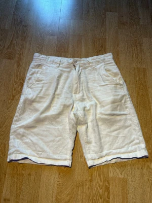 Vita linneshorts med fickor - Säljer ett par stilrena vita shorts i linne med normal passform. Shortsen har sidofickor fram, bakfickor med knapp och stängs med knapp och dragkedja. Perfekta för varma sommardagar och ger en fräsch look.