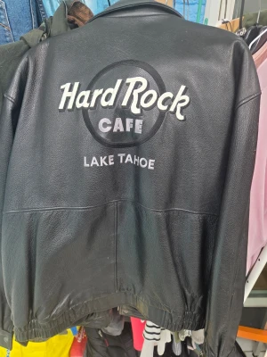 Hard Rock Cafe svart skinnjacka - Svart skinnjacka från Hard Rock Cafe med stort broderat motiv på ryggen där det står 'Hard Rock Cafe Lake Tahoe'. Klassisk modell med bred krage, dragkedja och resår i midjan. Perfekt för dig som gillar ikoniska plagg och vill sticka ut. Mktvbra skick inga hålnmm