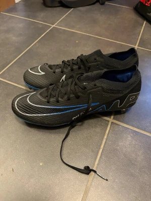 Nike Mercurial svarta fotbollsskor - Säljer ett par Nike Mercurial fotbollsskor i svart med vita och blå detaljer. Skorna har snörning, låg profil och är tillverkade i ett lätt syntetmaterial för bra bollkontroll och grepp på planen. Perfekta för dig som vill ha snabbhet och stil på fotbollsplanen.