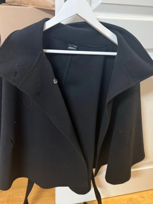Svart poncho/cape från Gina Tricot - Säljer en superfin svart poncho/cape från Gina Tricot. Väldigt stilren och passar perfekt över outfits på hösten/vintern 🍂✨Den har ett snyggt fall och knytning i midjan som gör att den sitter jättefint. Enkel att styla både casual och mer uppklätt 🫶 Skriv gärna om du vill ha fler bilder eller mått 💌