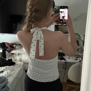 Vit prickig halterneck topp - Säljer en söt vit halterneck topp med svarta prickar och knyt i nacken. Toppen är ärmlös, har smockad resår i midjan och en lätt v-ringad front. Perfekt för dig som gillar en feminin och trendig stil.