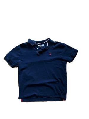 Marinblå pikétröja Assn Polo - Snygg marinblå pikétröja från polo assn med klassisk krage och knappar framtill. Tröjan har korta ärmar. Liten broderad logga på bröstet ger en stilren touch. OBS, det är inte ralph lauren utan polo assn.