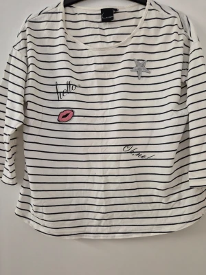 Tröja - Vit långärmad tröja från Kappahl med tunna svarta ränder och coola broderade detaljer som stjärna, läppar och texten 'hello' och 'oh no!'. Tröjan har rund hals och är gjord i mjukt bomullsmaterial. Perfekt för en avslappnad och trendig look. Finns hål och lite gult underarmarna.
