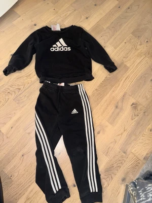 Svart Adidas träningsset med vita ränder - Snyggt svart träningsset från Adidas med matchande sweatshirt och byxor. Tröjan har stor Adidas-logga på bröstet och byxorna har de klassiska tre vita ränderna längs benen samt Adidas-logga. Perfekt för sport eller chill. Materialet är mjukt och bekvämt.Svart Adidas träningsset med vita ränder