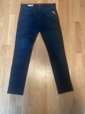 Mörkblå slim fit jeans från Replay - Snygga mörkblå slim fit jeans från Replay med klassisk femficksdesign och diskreta kontrastsömmar. Jeansen har en rak passform och detaljerad logotyp på myntfickan. Perfekta för en trendig och avslappnad stil.