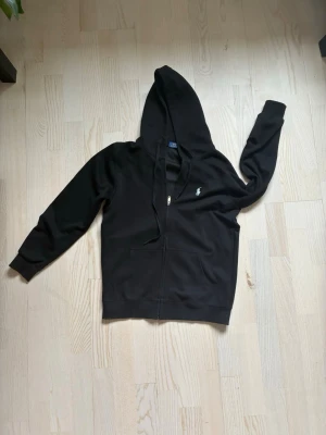 Svart hoodie från Polo Ralph Lauren - Svart hoodie med dragkedja från Polo Ralph Lauren. Klassisk design med vit broderad logga på bröstet. Tillverkad i mjukt bomullsmaterial som är skönt att ha på sig.