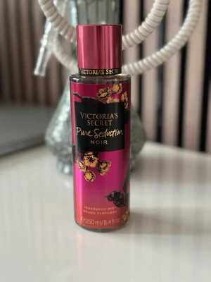 Victoria's Secret Pure Seduction Noir - En snygg parfymmist från Victoria's Secret i doften Pure Seduction Noir. Flaskan är metallicrosa med svarta och guldiga detaljer samt blommotiv. Rymmer 250 ml och har en lyxig känsla, perfekt för dig som gillar fruktiga och blommiga dofter.