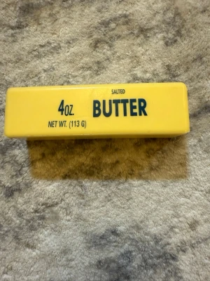 Butter Squishy - En gul rektangulär plastbit med texten '4oz BUTTER SALTED' tryckt i svart. Ser ut som ett smörpaket men är tillverkad i plast, troligen som dekoration eller leksak. Måtten anges till 113 gram.