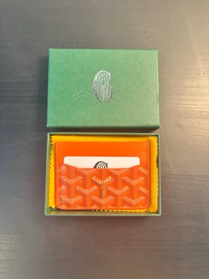 Orange kortfodral  - Snyggt orange kortfodral med det klassiska mönstret i vitt och svart. Tillverkat i canvas med läderdetaljer. Kommer i original grön ask med logga. Perfekt för kort och ID.