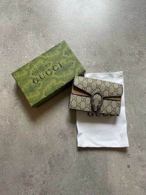  Dionysus GG Supreme handväska - Säljer en handväska i beige GG Supreme canvas med klassiskt monogrammönster och silverfärgat spänne med tigerhuvud. Väskan har en kompakt, rektangulär form och detaljer i brunt läder. Kommer med originalkartong och dustbag.