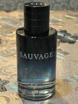 Dior Sauvage Eau de Toilette - Dior Sauvage Eau de Toilette i en elegant cylinderformad glasflaska med svart och blå toning. Flaskan har tydlig logga och ett stilrent svart lock med diskret Dior-märkning på toppen. En ikonisk doft som utstrålar lyx och modernitet.