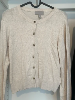 Beige kofta från H&M med knappar - Klassisk beige kofta från H&M med rund halsringning och långa ärmar. Koftan har ribbade muddar och knäpps med sex stilrena knappar framtill. Perfekt att slänga över en t-shirt eller linne för en enkel och clean look.