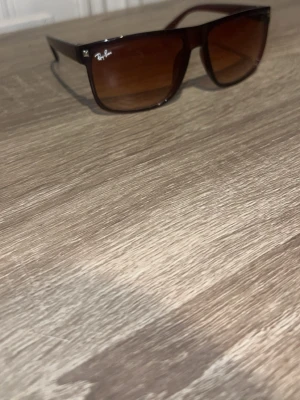 Bruna Ray-Ban solglasögon - Säljer ett par stilrena solglasögon från Ray-Ban med bruna, fyrkantiga bågar och matchande bruna glas. Klassisk logga på linsen och diskreta metall-detaljer vid hörnen. Perfekta för soliga dagar och ger en cool vibe.
