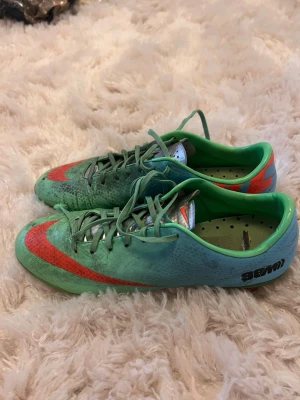 Nike Mercurial fotbollsskor grön/röd - Säljer ett par Nike Mercurial fotbollsskor i grön färg med röda detaljer och svart logga på hälen. Skorna har låg profil, snörning och ovandel i syntetmaterial för bra bollkontroll. Perfekta för dig som vill sticka ut på planen!
