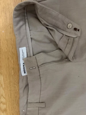 Beige kostymbyxa från Bershka - Snygga beige kostymbyxor från Bershka med raka ben och hög midja. Byxorna har bälteshällor, dold dragkedja och knappstängning framtill. Tillverkade i ett mjukt och lätt tyg som ger en clean och stilren look.