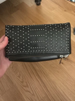 Svart kuvertväska med nitar - Snygg svart kuvertväska i läderimitation med silvriga nitar i ett rutmönster. Väskan har dragkedja och stilren design som ger en edgy vibe. Perfekt accessoar för att lyfta din outfit.
