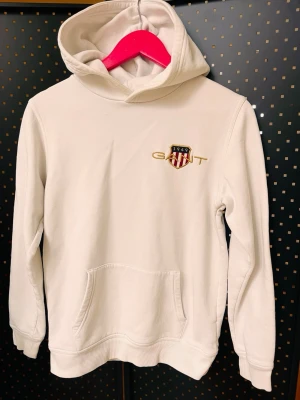 Vit hoodie från GANT med broderad logga stl 158/164 - Vit hoodie från GANT med broderad logga och sköld på bröstet. Klassisk känguruficka framtill och huva. Mjuk bomullskänsla och långärmad modell, perfekt för en chill och stilren look.