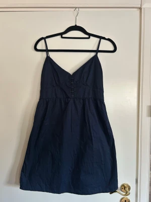 Marinblå klänning från Brandy Melville - Söt marinblå kortklänning från Brandy Melville med smala axelband och knappar framtill. Klänningen har en v-ringad urringning och är tillverkad i ett lätt bomullstyg, perfekt för varma dagar.