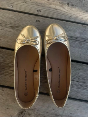 Guldiga ballerinaskor med rosett - Snygga ballerinaskor i metallic guld från Claudia Ghizzani. Skorna har rund tå och en söt rosett framtill. Tillverkade i ett glansigt syntetmaterial som ger en trendig look. Endast använda vid ett tillfälle