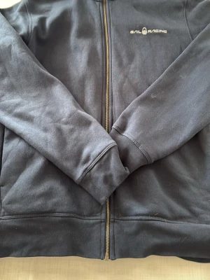 Mörkblå zip-hoodie från Sail Racing - Mörkblå hoodie med dragkedja från Sail Racing. Klassisk modell med ribbade muddar och fickor på sidorna. Diskret logga på bröstet. Perfekt för en avslappnad och sportig stil.