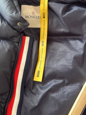 Mörkblå dunväst från Moncler Genius - Snygg mörkblå dunväst från Moncler Genius med klassisk röd, vit och blå rand vid dragkedjan. Västen har en gul detalj med Moncler Genius-tryck och ett unikt serietidningsmärke på insidan. Perfekt för dig som gillar exklusiva och trendiga plagg.