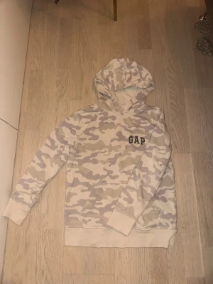 Beige camo hoodie från GAP - Säljer en vit och ljusgrå camo-mönstrad hoodie från GAP med huva och ribbade muddar. GAP-loggan är tryckt på bröstet. Perfekt för dig som gillar streetwear och avslappnad stil.