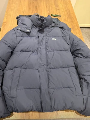 Mörkblå pufferjacka från Calvin Klein - Snygg mörkblå pufferjacka från Calvin Klein med avtagbar huva och dragkedja framtill. Jackan har CK-logga broderad på bröstet och stora quiltade partier för extra värme. Perfekt för kalla dagar och riktigt trendig look.