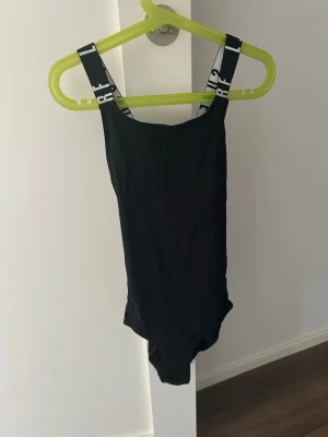 Svart baddräkt med breda axelband - Snygg svart baddräkt med raka linjer och breda axelband med vit textdetalj. Axelbanden ger en sportig känsla och baddräkten har en klassisk, enkel design. Perfekt för stranden eller poolen.