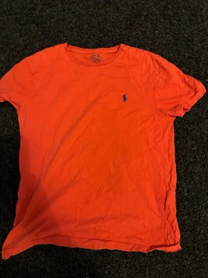Röd/orange Polo Ralph T-shirt  - Snygg röd t-shirt från Polo Ralph Lauren med klassisk blå logga broderad på bröstet. T-shirten har rund halsringning och korta ärmar. Tillverkad i mjuk bomull för en bekväm känsla. .