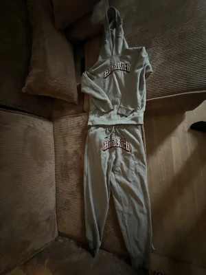 Grått loungewear-set från MindRich - Mysigt grått loungewear-set från MindRich med matchande hoodie och byxor. Hoodien har stor ficka framtill och tryckt logga på bröstet, medan byxorna har resår i midjan och logga på ena benet. Perfekt för chill dagar hemma.