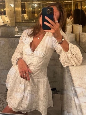 Vit kort klänning med spetsdetaljer - Söt vit kortklänning med v-ringning, långa ärmar och fina spetsdetaljer. Klänningen har volang nertill och markerad midja som ger en feminin siluett. Perfekt för sommarens alla tillfällen och har en romantisk känsla.