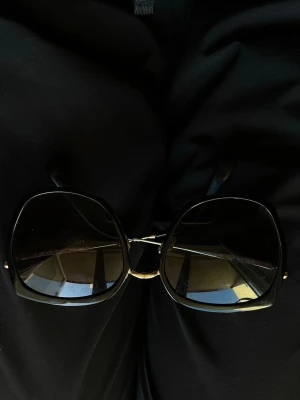 Svarta solglasögon med fyrkantig form - Snygga solglasögon med stora, fyrkantiga svarta bågar och mörka glas. Metallbrygga i guld som ger en cool kontrast. Perfekt för dig som vill ha en trendig och stilren look. Passar till sommarens alla outfits.