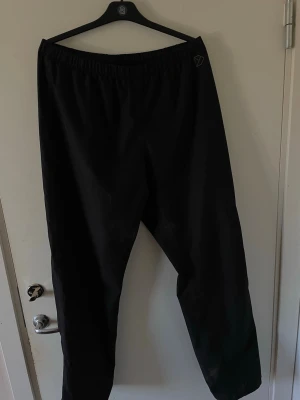 Svarta mjukisbyxor från didriksons - Säljer ett par svarta byxor med elastisk midja och diskret logga på höften. Byxorna har en avslappnad passform och är perfekta för träning. Materialet känns luftigt och bekvämt.
