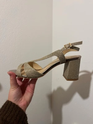 Glittriga sandaler med blockklack - Säljer ett par glittriga sandaler med bred blockklack och öppen tå. Remmarna är korslagda framtill och har ett justerbart spänne vid ankeln. Färgen är champagne/guld och materialet skimrar snyggt. Perfekta till bal eller student