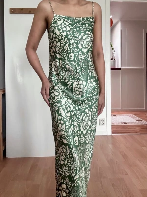 Grön blommig långklänning från Zara - Supersnygg långklänning från Zara i grönt och vitt med blommigt mönster. Klänningen har ärmlös design och figurnära passform som ger en elegant siluett. Perfekt för dig som vill sticka ut med ett unikt print och fräsch färg.