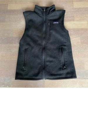 Svart fleeceväst från Patagonia - Stilren svart fleeceväst från Patagonia med hög krage och hel dragkedja framtill. Västen har två sidofickor med dragkedja och en bröstficka med dragkedja. Perfekt att slänga över en hoodie eller tröja för extra värme.