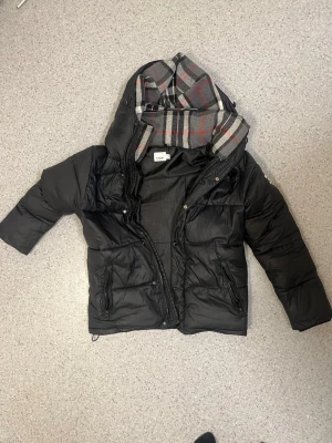 PROBLEM= problemen med jackan kan man se på bild 4 och att dragkedjan är ganska trög/ Svart dunjacka från Burberry - Snygg svart dunjacka från Burberry med klassiskt rutigt foder i huvan. Jackan har dragkedja, tryckknappar och två fickor framtill. Perfekt för kalla vinterdagar och har en modern, puffig passform.