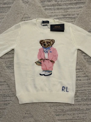 Vit stickad tröja Polo Ralph Lauren - Vit stickad tröja från Polo Ralph Lauren med ikonisk björn i rosa kostym på framsidan och RL-broderi nedtill. Tröjan har rund halsringning och ribbade muddar vid ärmslut och nederkant. Perfekt för dig som gillar klassisk preppy stil.