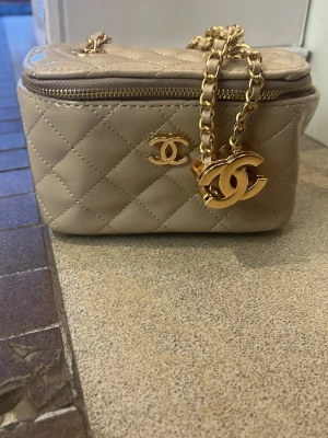 Beige axelremsväska med pärldetaljer - Snygg beige handväska i krokodilpräglat material med silverfärgad kedja och ett stort spänne dekorerat med pärlor och glittrande stenar. Väskan har en stilren, rektangulär form och passar perfekt för dig som gillar trendiga accessoarer.